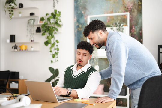Two men collaborate on a laptop in a modern office space, one actively typing while the other leans in to offer guidance and discussion.
