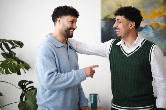 Two young men share a laugh and a friendly gesture indoors, one with his arm around the other's shoulder.