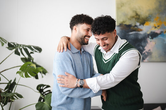 Two men share a joyful moment, embracing and laughing together in a bright, modern interior with a large plant and abstract art.
