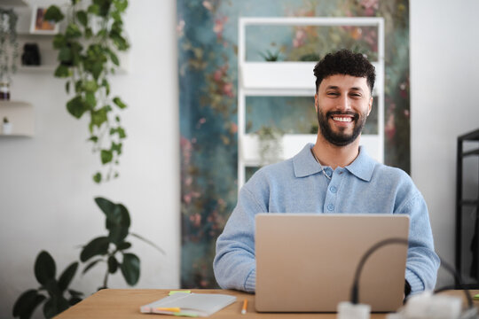 A smiling man sits at a desk with a laptop, working in a bright, plant-filled room.