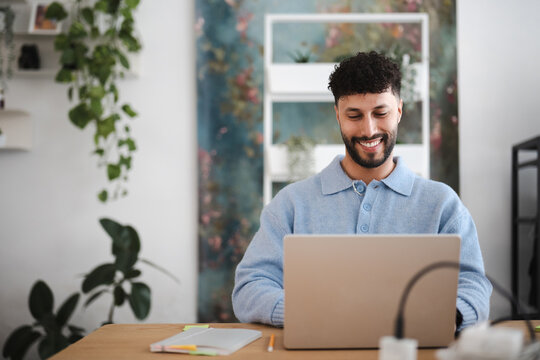 A man smiles while working on his laptop in a bright, plant-filled room. He is dressed in a light blue sweater and sits at a wooden desk.