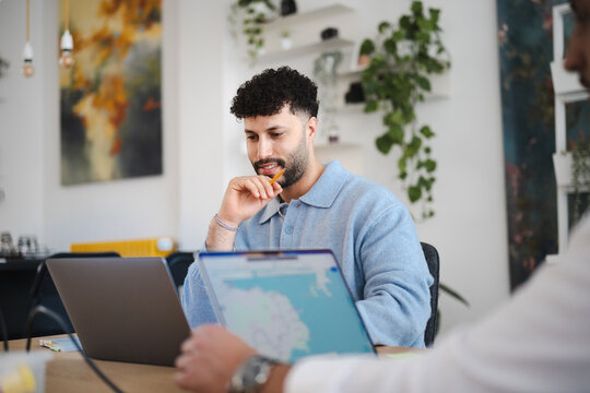 A man with curly hair sits at a desk, holding a pencil to his chin as he looks at a laptop screen. He is in a modern office space with plants and artwork.