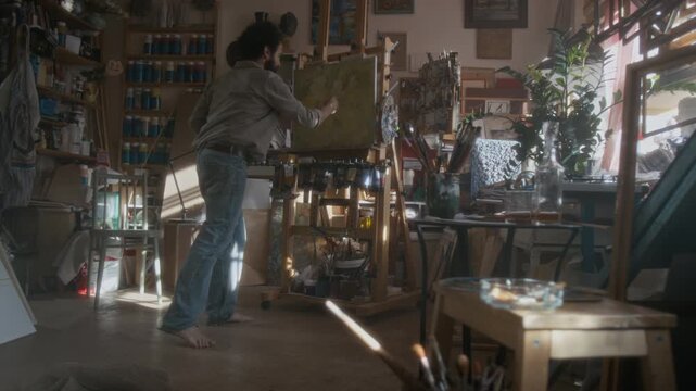 Full length shot of nervous male artist drawing at easel in cluttered studio, throwing and kicking canvas during mental breakdown, then sitting on floor to continue his work