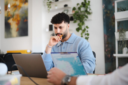 A man with curly hair sits at a desk, looking intently at a laptop screen while holding a pencil to his chin. He is in a modern office space with plants and artwork.