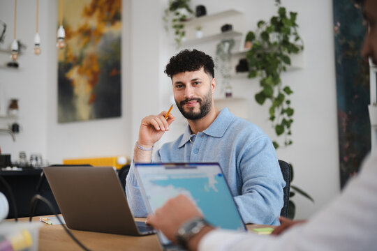A man with curly hair holds a pencil while looking at a laptop in a modern office setting. He is smiling and appears engaged in his work.