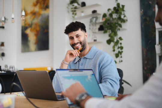 A man smiles while holding a pencil, working on a laptop with another person in a modern office setting.