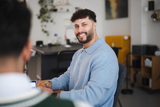A man with curly hair smiles while sitting at a desk with a pencil and tablet, engaged in a discussion with another person.