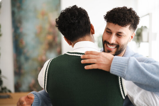 Two men embrace warmly, one with a joyful smile, sharing a moment of connection and affection in a bright, modern setting.