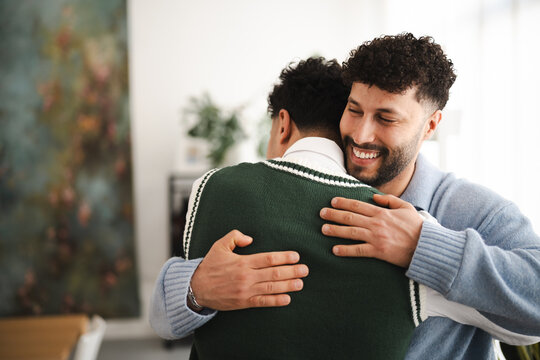 Two men embrace warmly indoors, one with a wide smile and closed eyes, sharing a moment of connection and affection.