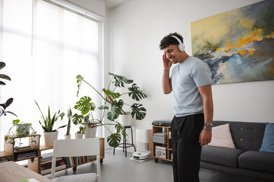 A young man with curly hair wearing headphones smiles while listening to music in a bright, modern living room with plants and artwork.