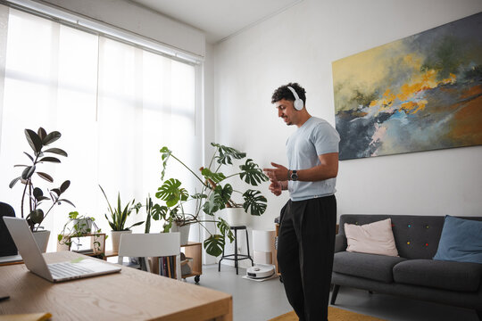A man wearing headphones dances in his living room. He is surrounded by plants and a laptop on a desk.