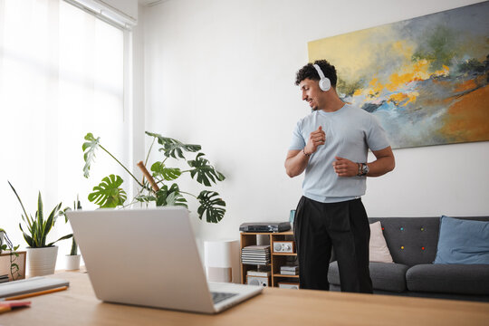 A young man wearing headphones dances joyfully in his bright, modern living room while listening to music.