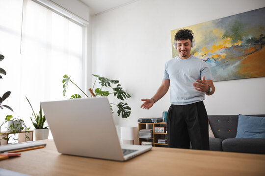 A young man wearing headphones gestures while looking at his laptop, engaged in a video call or online presentation in a bright, modern room.