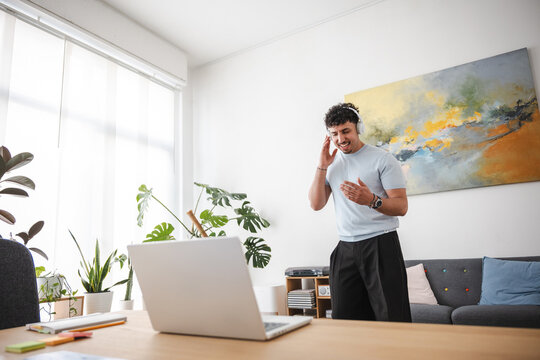 A young man wearing headphones is enthusiastically singing along to music while working on his laptop in a bright, modern home office.
