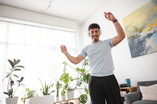 A young man with curly hair wears headphones and dances joyfully in a bright room filled with plants.
