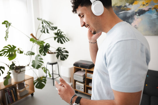 A young man with curly hair wears headphones and smiles while listening to music in a bright room with plants and artwork.