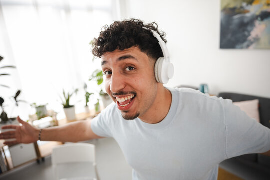 A young man with curly hair wears headphones and smiles broadly, gesturing with his hands while listening to music in a bright, modern room.