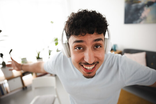 A young man with curly hair wears headphones and smiles broadly, his arms outstretched in a gesture of joy and excitement while listening to music.