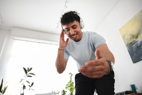 A young man with curly hair wears headphones and smiles while gesturing with his hand, enjoying music in a bright room with plants.
