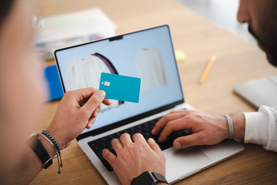 A person holds a payment card while another person types on a laptop, suggesting online shopping or a financial transaction.