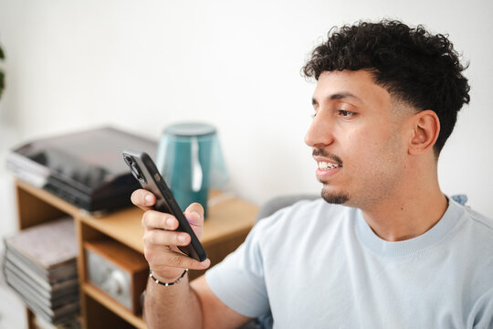 A young man with curly hair smiles while looking at his smartphone. He is holding the device in his hand, engaged with its screen.