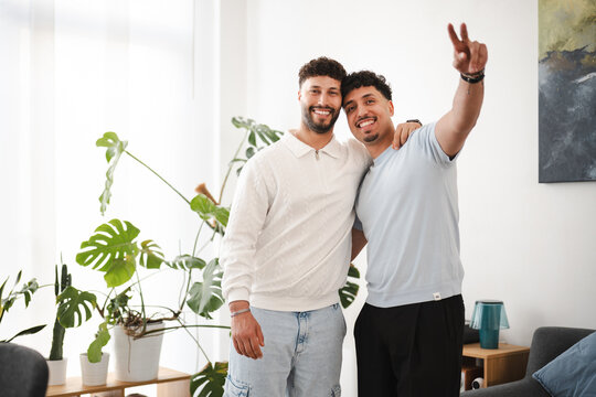 Two smiling men embrace in a bright room with plants. One man holds up two fingers in a peace sign.