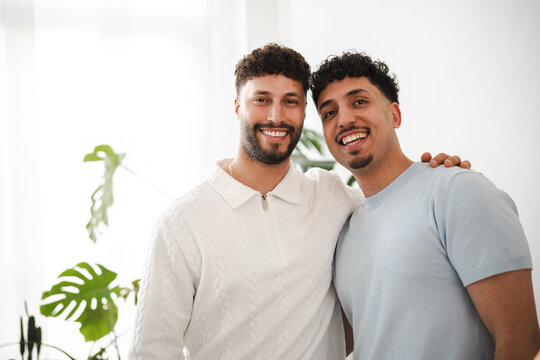 Two smiling men stand together, one with his arm around the other's shoulder, in a bright, indoor setting with a large plant in the background.