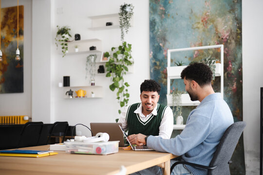 Two men collaborate on a laptop at a wooden table in a bright, modern office space with plants and artwork.