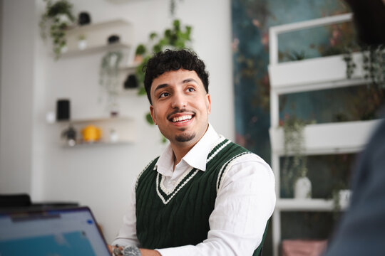 A young man in a white shirt and green sweater vest smiles while looking away from the camera during a meeting.