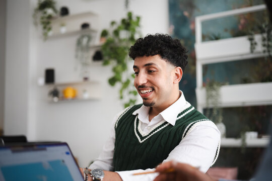 A young man smiles while working on a laptop in a modern office setting with plants.
