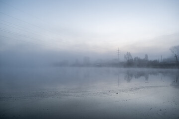 Fototapeta premium Fog envelops a tranquil lake at dawn, with reflections of trees and power lines visible on the water's surface enhancing the serene scene