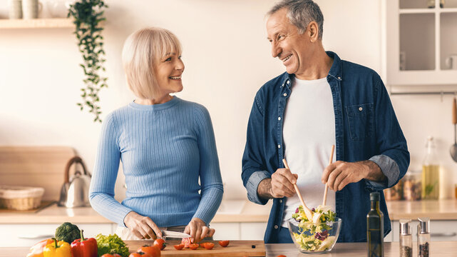 A man and a woman are preparing a meal in a bright kitchen. The woman is chopping vegetables while the man mixes a salad. They are happy and enjoying their time together.