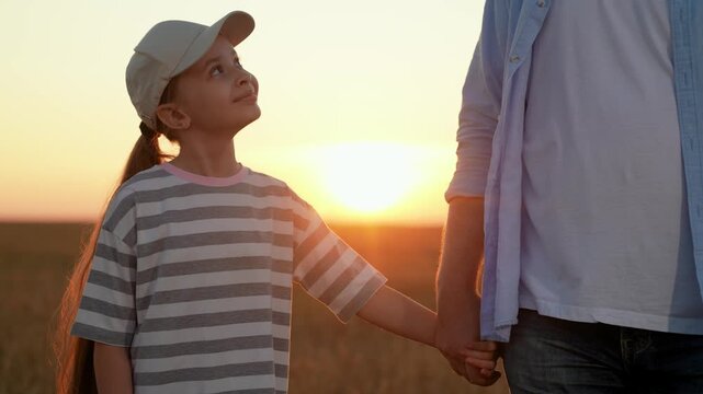 Dad walks with little daughter, holding hands, in field sunset. Family picnic in park. Happy family enjoys outdoor adventure. Smiling child holds daddy hand. Father Day. Dad raises daughter. Kid girl