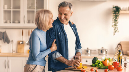 In a bright kitchen, a couple stands close while preparing food. The man is chopping vegetables on a cutting board. The woman smiles and leans against him. © Prostock-studio