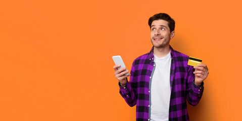 A young man stands against an orange wall. He holds a smartphone in one hand and a credit card in the other hand. He appears to be thinking about shopping or making a purchase. © Prostock-studio