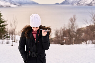 A woman stands in the snow, speaking on her phone. She is near a lake with snow-covered mountains in the background. The sun sets in the distance, giving a warm glow. © Eclipsnet