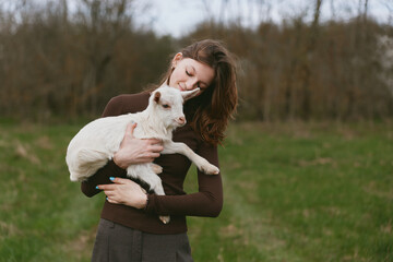 Obraz premium A woman in a brown sweater cradles a white goat kid in a sunlit meadow, smiling softly as they share a quiet moment. A lifestyle scene of animal bonding and care.