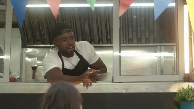 Wide camera view of African American male speaking with customer at window. Man leaning from food truck. Confirming order with hand gesture. Wearing apron and cap. Busy outdoor food stand scene.