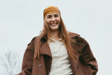 Obraz premium Cheerful woman outdoors, smiling, wearing a warm brown coat and bandana, casual pose, natural colors, soft breeze, sunny atmosphere, candid lifestyle portrait, autumn tones, relaxed vibe, simple