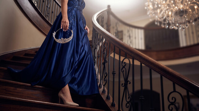 Woman in blue gown descending staircase while holding a tiara  