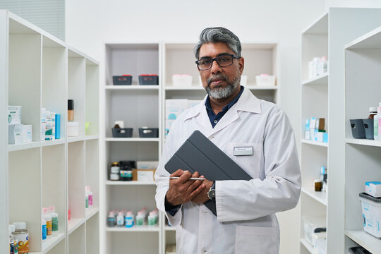 Portrait of middle aged Indian male pharmacist holding tablet and stylus in pharmacy, managing inventory and digital records. For healthcare retail, medication services, telehealth marketing