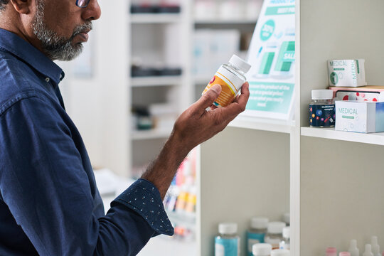 Middle aged man examining supplement bottle in pharmacy aisle, comparing product information for purchase decision. Useful for healthcare retail, medication choice, consumer wellness