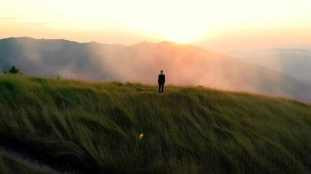 long shot of a person standing alone on a grassy hill surrounded by thick mist and fog during a golden sunrise