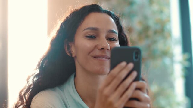 Woman with medium skin tone and long curly hair looks at her phone with a smile. She sits inside a well-lit room surrounded by plants, enjoying her time on the device.