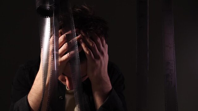 Person covering face with hands showing signs of frustration and stress, with film negatives hanging around embodying the concept of creative block and memory loss in a dark, cinematic environment
