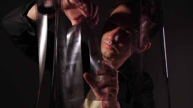 Filmmaker holding and carefully inspecting a celluloid film strip against the light in a darkroom or editing studio, reviewing frames for production or post production work