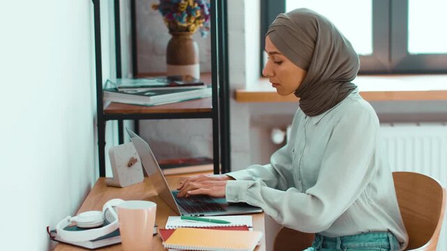 A woman with light skin sits at a desk in a bright room, focused on her laptop. She has a neutral expression and is surrounded by notebooks and a cup, showing a dedicated work environment.
