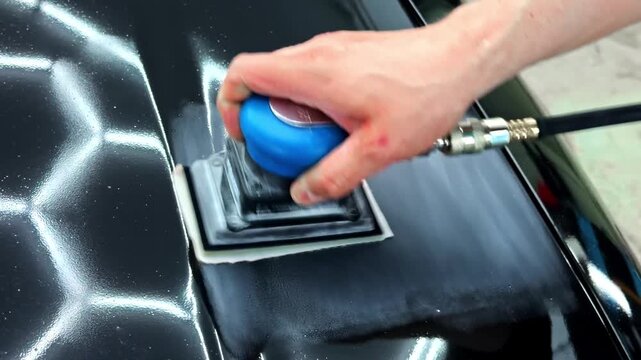 Close-up of a hand using a pneumatic sanding block on a black car panel, showing paint correction, body preparation, and professional auto detailing work.