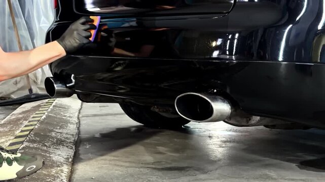 Worker applies detailing product to car rear bumper in indoor garage setting