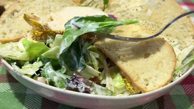 Caesar salad served with large pieces of croutons and grilled chicken, topped with an edible flower in the center. dish is presented on a red, green, and white checkered tablecloth in a rustic style.